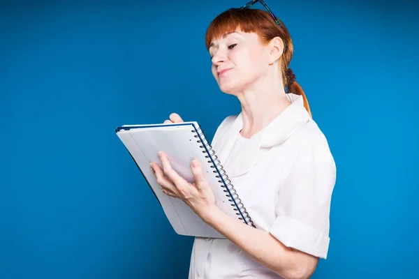 Rothaariges Mädchen in medizinischer Kleidung schreibt in ein Notizbuch einer Platte, isoliert auf blauem Hintergrund — Stockfoto