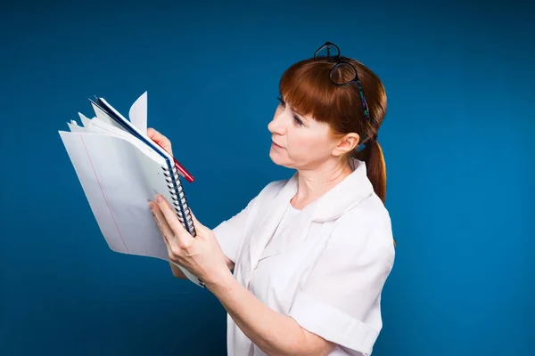 Rothaariges Mädchen in medizinischer Kleidung schreibt in ein Notizbuch einer Platte, isoliert auf blauem Hintergrund — Stockfoto