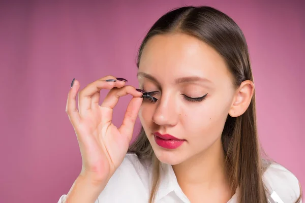 Menina bonita experimenta pestanas falsas, em um fundo rosa, isolado — Fotografia de Stock