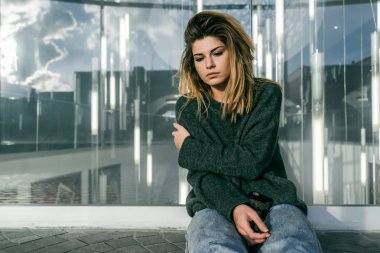 üzgün sarışın saçlı genç kız bir cam bina önünde oturur