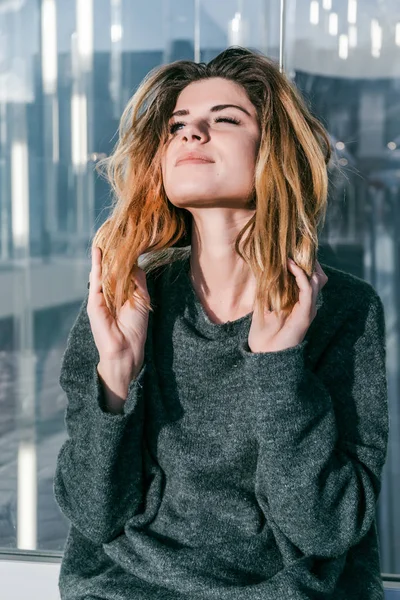 Das Mädchen sitzt an der Fassade eines Glasgebäudes und spielt mit den Haaren — Stockfoto