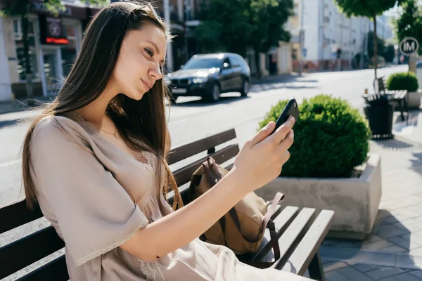 Linda chica sentada en un banco en una calle de la ciudad y haciendo selfie — Foto de Stock