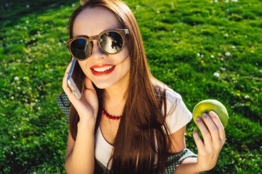 Mutlu gülümseyen kadın güneş gözlüğü, cep telefonunu kullanma ve elma tutar