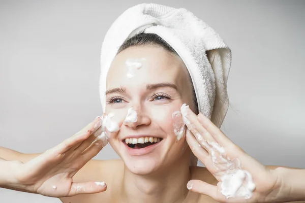 Mujer divertida en una toalla en la cabeza feliz limpia la piel con espuma sobre un fondo blanco aislado. Concepto de limpieza del cuidado de la piel —  Fotos de Stock