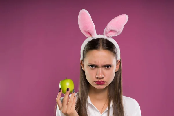 Egy dühös lány rózsaszín nyúl füle almát tart a kezében — Stock Fotó
