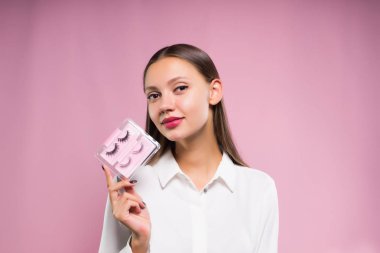 Kız bir gömlek pembe bir arka plan üzerinde sahte kirpik bir kümesini onun elinde tutar.