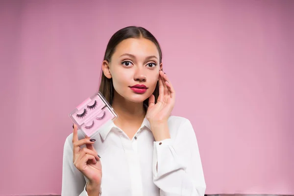 Belle fille avec de gros yeux regarde la caméra et tient dans les mains faux cils, sur un fond rose — Photo