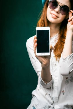 Kızıl saçlı gülümseyen kız bir şey telefonun ekranında gösterilen ve kameraya bakarak büyük yuvarlak güneş gözlüğü