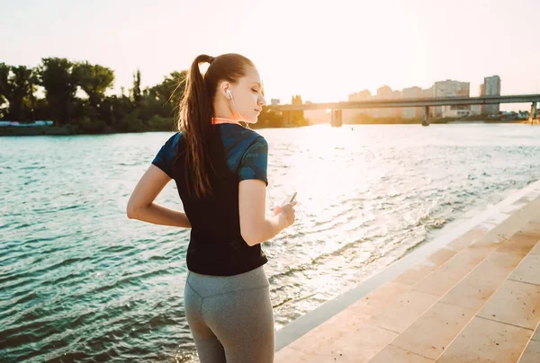 Flicka i solnedgången på en joggingtur lyssnar på musik i hörlurar — Stockfoto