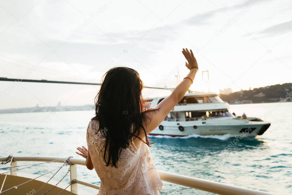 Young girl traveler relaxes during a cruise on a yacht, waving her hand to a passing ship at sunset, a bridge against a background