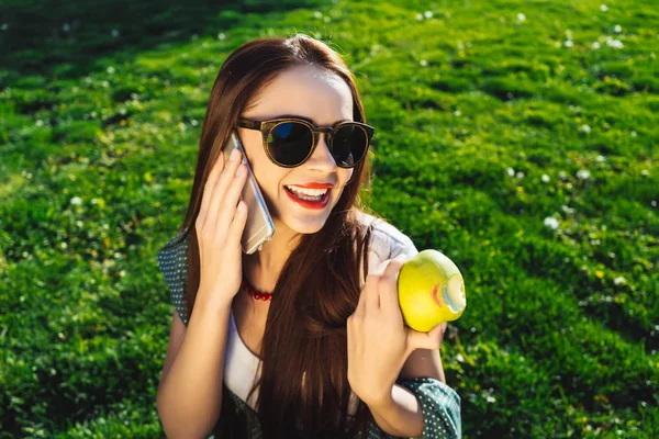 Talking woman holds mobile,sitting on grass in dress,sunglasses