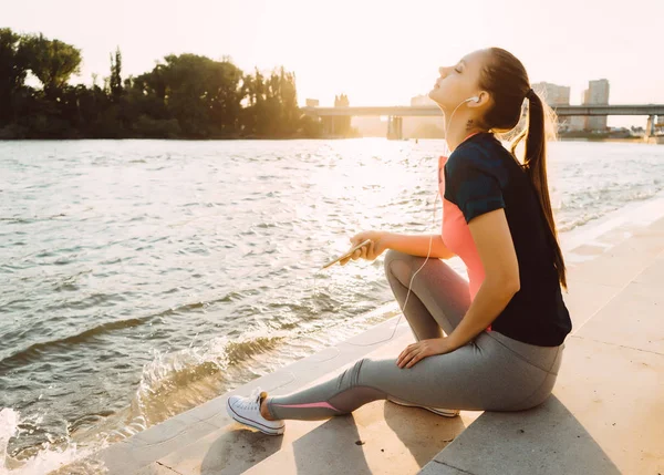 Dziewczyna po joggingu siedzi na brzegu rzeki o zachodzie słońca i słucha muzyki — Zdjęcie stockowe