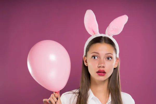 Una ragazza con le orecchie divertenti sulla testa tiene una palla rosa tra le mani e guarda la fotocamera in confusione — Foto Stock