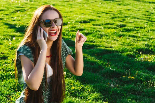 Happy woman in glasses pointing to something on grass, uses mobile — Stock Photo, Image