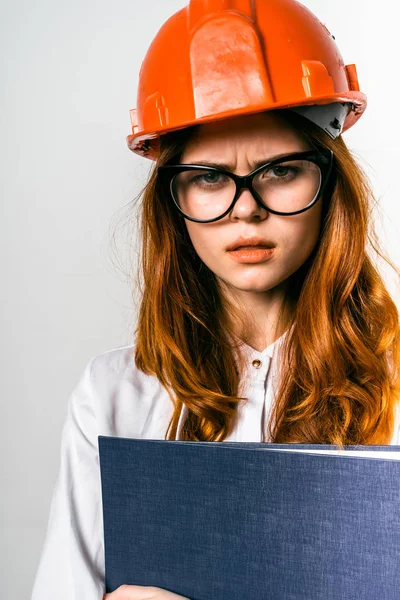 Uma menina com raiva em um capacete de construção laranja está segurando uma pasta — Fotografia de Stock