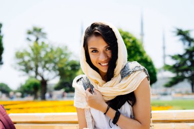 Müslüman kadının bir atkı ve beyaz elbise bir caminin arka planda bir bankta oturan Modern genç gülümseyen