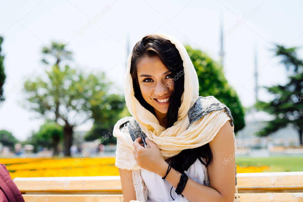 Modern young Muslim woman in a kerchief and white dress is sitting on a bench in the background of a mosque, smiling