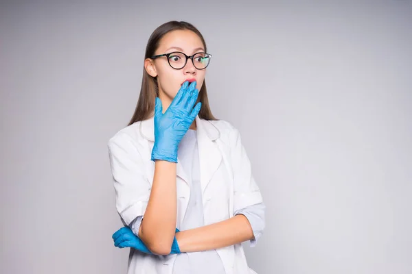 En flicka i en medicinsk klänning och handskar, täcker munnen med överraskning — Stockfoto