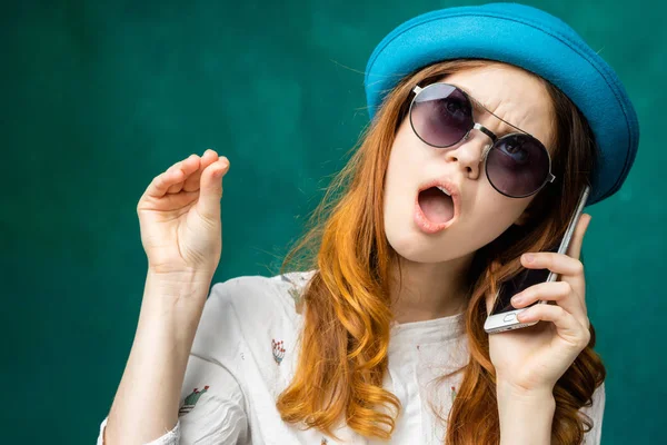 Рыжая девушка в очках и в синей шляпе разговаривает по телефону с плохим настроением — стоковое фото