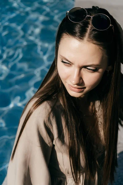 Menina em óculos de sol senta-se à beira da água azul ao sol, retrato — Fotografia de Stock