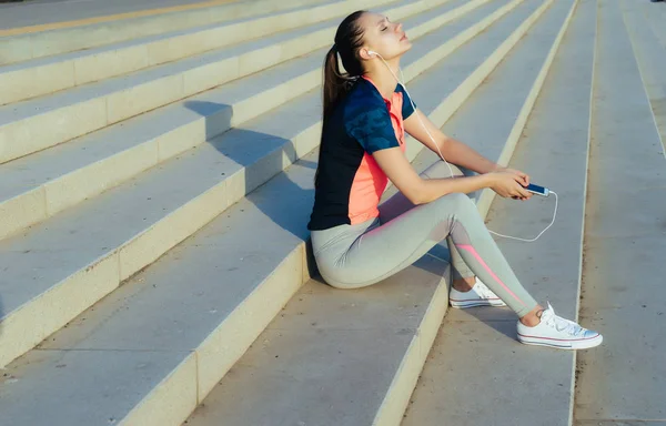 Een meisje in sportkleding is rustend op de trap en luisteren naar muziek op koptelefoon — Stockfoto