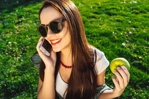 La chica está sentada en el césped, hablando en el teléfono inteligente y riendo. sostiene una manzana en su mano — Foto de Stock