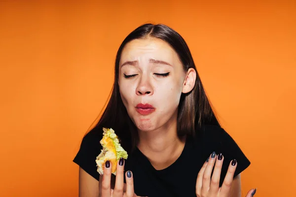 Ein frustriertes Mädchen beißt aus ihrem Burger auf orangefarbenem Hintergrund — Stockfoto