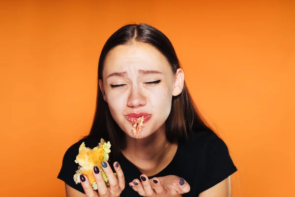 Hayal kırıklığına uğramış kız turuncu bir arka plan üzerinde onun elinde bir hamburger yiyor — Stok fotoğraf
