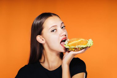 Aç kız hevesle turuncu bir arka plan üzerinde izole bir hamburger yiyor