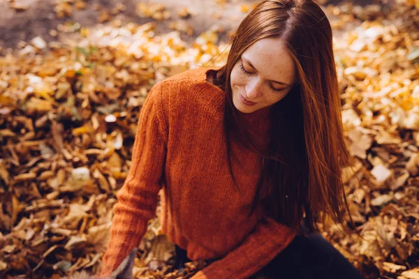 Un otoño dorado, una chica encantadora se sienta en hojas en un suéter cálido y mira cuidadosamente hacia fuera, un ambiente acogedor — Foto de Stock