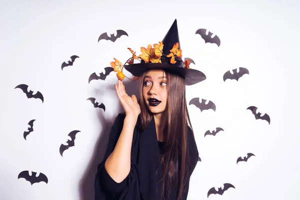 Schattig meisje heks gonna Halloweenfeest en straighten van haar herfst bladeren op haar zwarte vreselijke hoed. Tegen de achtergrond van een bos van vleermuizen — Stockfoto