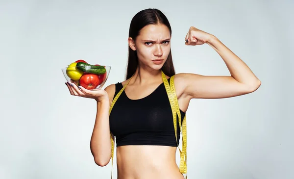 Sportovní dívka držící talíř zeleniny ve svých rukou a ukazuje její sílu, zdravý život — Stock fotografie