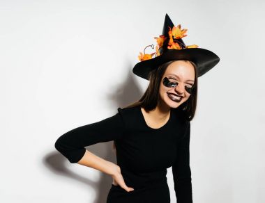 Siyah bir şapka ve gözlerinin altında siyah yamalar ile mutlu Halloween seksi cadı. Güzel genç kadın cadı şapka ve beyaz arka plan üzerinde Göl kenarında gülüyor kostüm