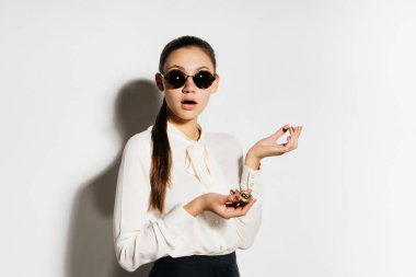 siyah gözlüklü Cool ciddi kız bir bozuk para tutar ve beyaz bir arka plan üzerinde izole kameraya benziyor