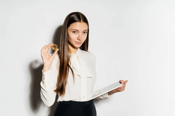Офисная девушка из банка смотрит в камеру и держит в руке какую-то бумагу и золотую монету, изолированную на белом фоне, криптовалюту, биткойны — стоковое фото