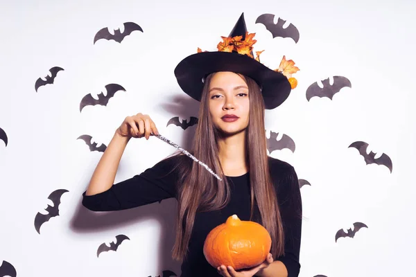 Happy Halloween Sexig häxa med en svart hatt och med en trollstav och en pumpa i händerna tittar in i kameran. Häxan ska en Halloweenfest. Isolerad mot bakgrund av fladdermöss — Stockfoto