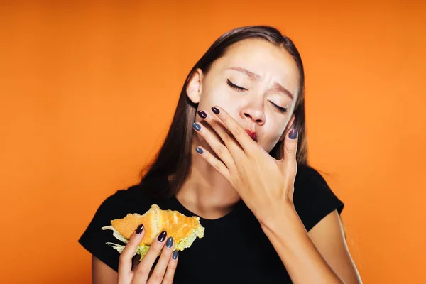 A rapariga esfomeada come o hambúrguer. A rapariga lambe os dedos. Comida muito saborosa, isolada em um fundo laranja — Fotografia de Stock