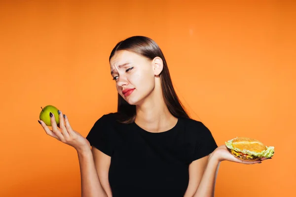 Dívka před těžkou volbu. Apple a hamburger. Lahodné jídlo nebo zdravých potravin. Izolované na oranžové pozadí — Stock fotografie