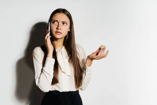 Uma garota indignada séria está falando ao telefone, segurando uma moeda em suas mãos e olhando para longe. Bitcoins, criptomoeda . — Fotografia de Stock