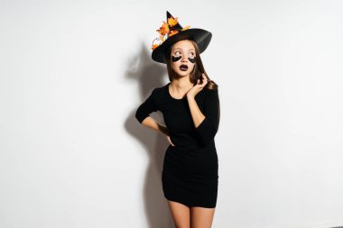 Siyah bir şapka ve gözlerinin altında siyah yamalar ile mutlu Halloween seksi cadı. Güzel genç kadın cadı şapka ve beyaz arka plan üzerinde Göl kenarında gülüyor kostüm