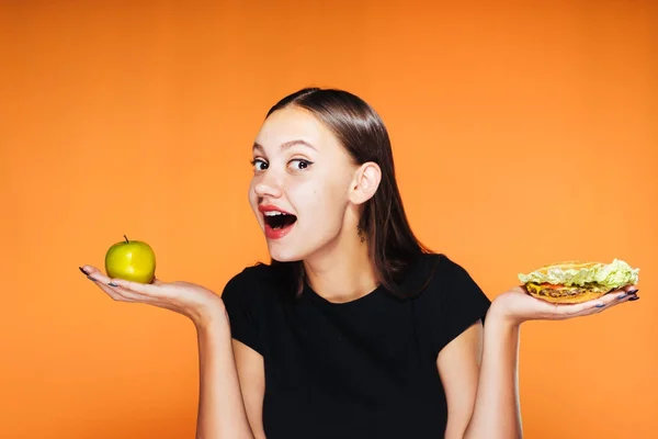 Uma menina triste escolhe que é melhor comer uma maçã ou um sanduíche com água na boca. comida saudável, comida deliciosa — Fotografia de Stock