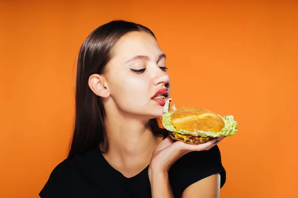 Menina faminta come ansiosamente um hambúrguer, isolado em um fundo laranja — Fotografia de Stock