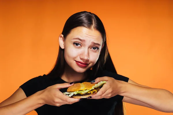 Мила дівчина тримає гамбургер в руках і дивиться на камеру, ізольована на помаранчевому фоні . — стокове фото
