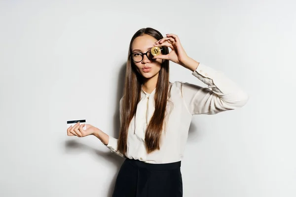 Uma menina bonita em uma blusa e usando óculos tem 2 moedas em suas mãos, e uma das moedas emoldurado-a ao olho. Bitcoins, criptomoeda — Fotografia de Stock