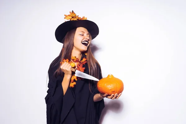 Śmiejąc się młoda dziewczyna ubrana w witch halloween, przytrzymaj dyni i nóż w ręce — Zdjęcie stockowe