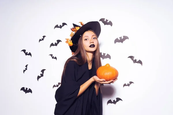 Una joven con un lápiz labial negro finge ser una bruja para Halloween, sostiene una calabaza naranja en su mano y se ve sombría — Foto de Stock