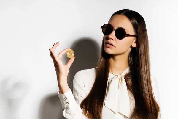 Uma empresária de cabelos escuros em óculos de sol está segurando um bitcoin de ouro, uma moeda criptomoeda — Fotografia de Stock