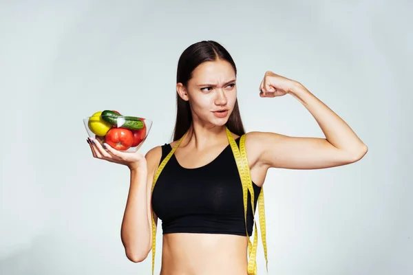 A jovem do esporte observa sua figura, segura um prato com legumes e frutas, mostra seu bíceps — Fotografia de Stock