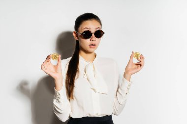 güneş gözlüklü genç sürpriz iş kadını altın bitcoins elinde tutar.