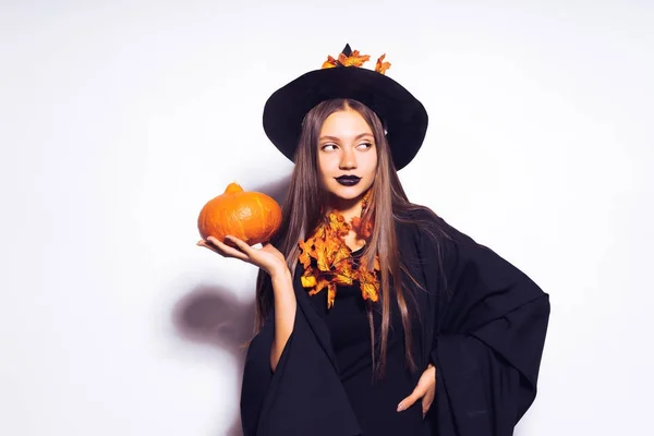 Jeune fille avec du rouge à lèvres noir sous la forme d'une sorcière pour Halloween, son manteau noir flottant, tenant une citrouille dans ses mains — Photo